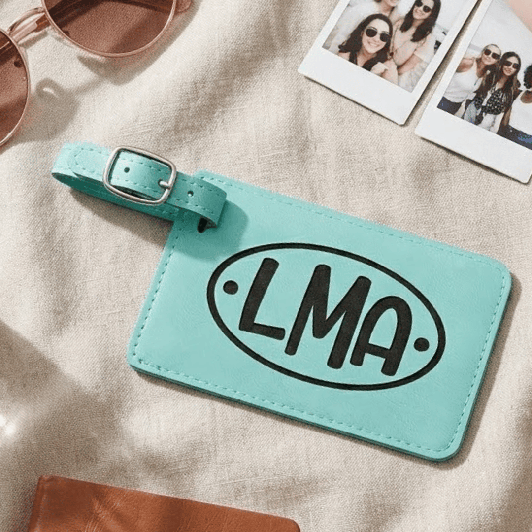Teal luggage tag with 'LMA' engraved in black on a beige surface with sunglasses and photos.