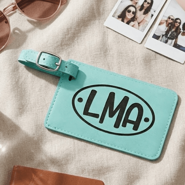 Teal luggage tag with 'LMA' engraved in black on a beige surface with sunglasses and photos.