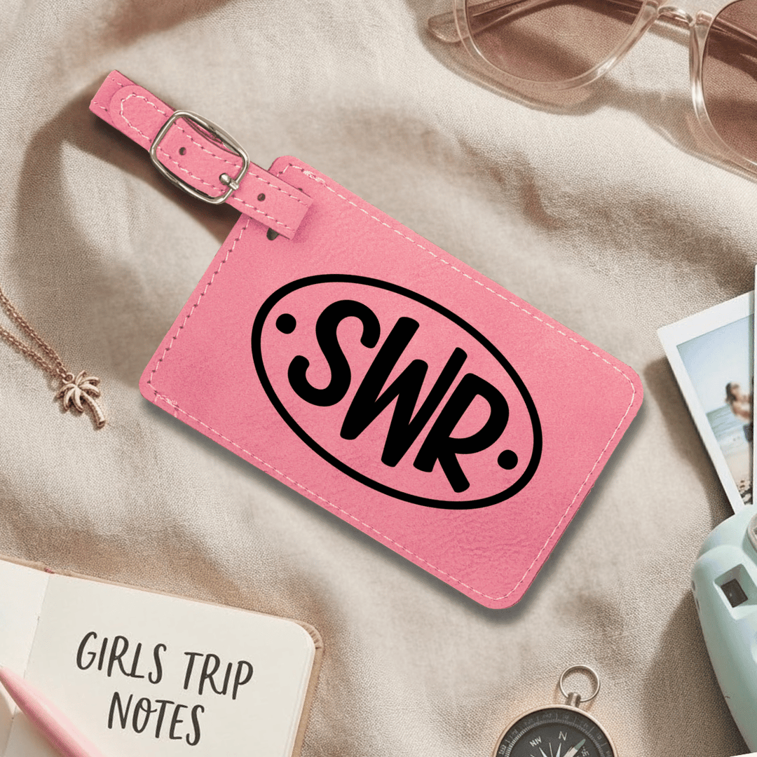 Pink luggage tag with 'SWR' initials engraved in black on a beige surface with travel items.