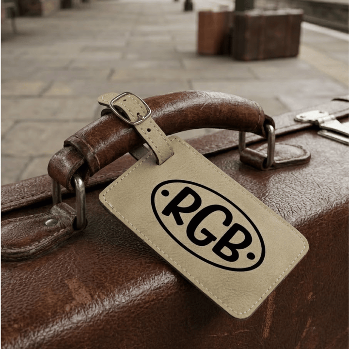 Brown leather suitcase with a beige tag featuring 'RGB' monogram engraved in black on a stone pavement background