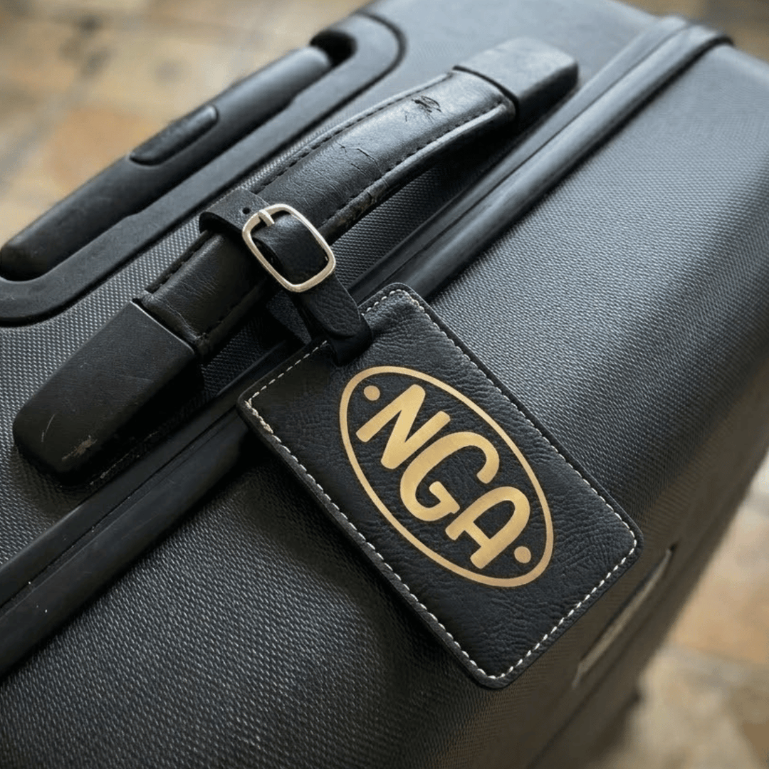 Black suitcase with a gold 'NGA' monogram engraving on a luggage tag, on a wooden surface.