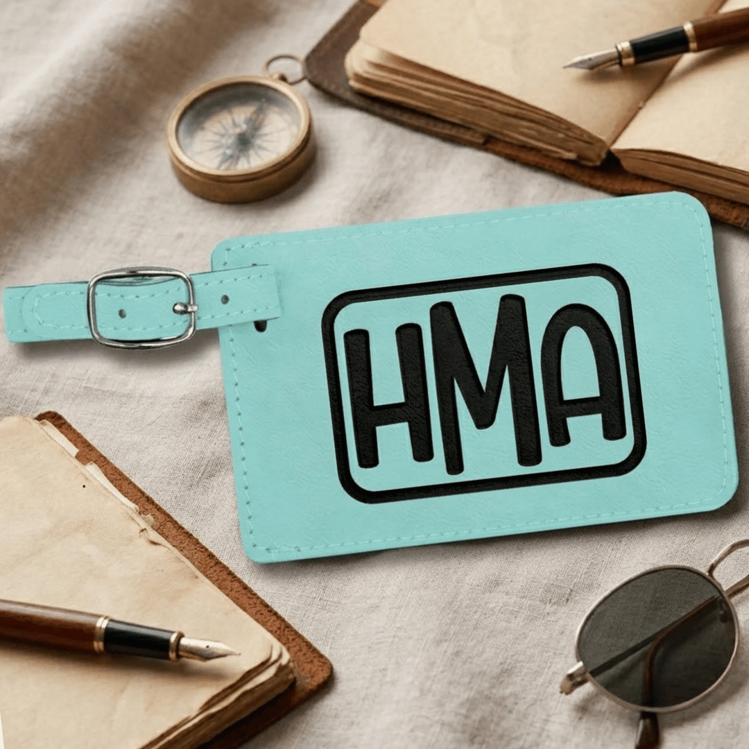 Teal luggage tag with 'HMA' engraved in black on tag, laying on a textured surface with books and sunglasses.