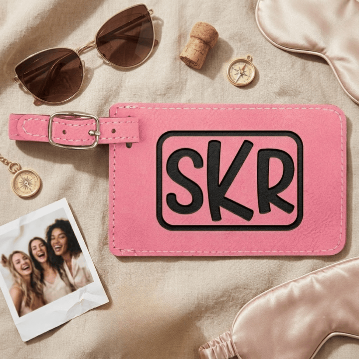 Pink luggage tag with 'SKR' engraved in a modern monogram laying on a beige surface with sunglasses, photo, and sleep mask.