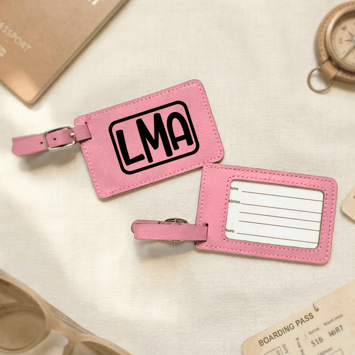 Front and back of pink luggage tag with 'LMA' engraved in black on front side and blank space for text on back, placed on a light surface with travel items.