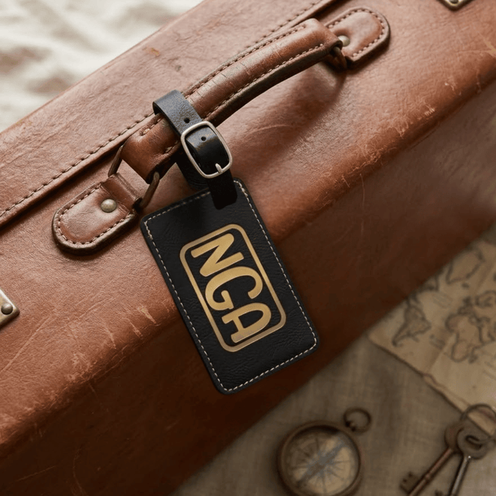 Brown leather suitcase with a black luggage tag featuring 'NCA' engraved in metallic gold on a textured surface.