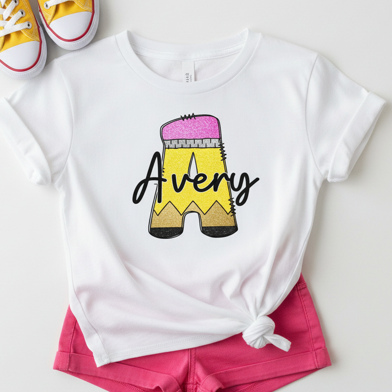 A white short-sleeve T-shirt laid flat with a tied hem, featuring a large pencil graphic and the name 'Avery' in black script. The shirt is styled with bright pink shorts and yellow sneakers on a light neutral background.