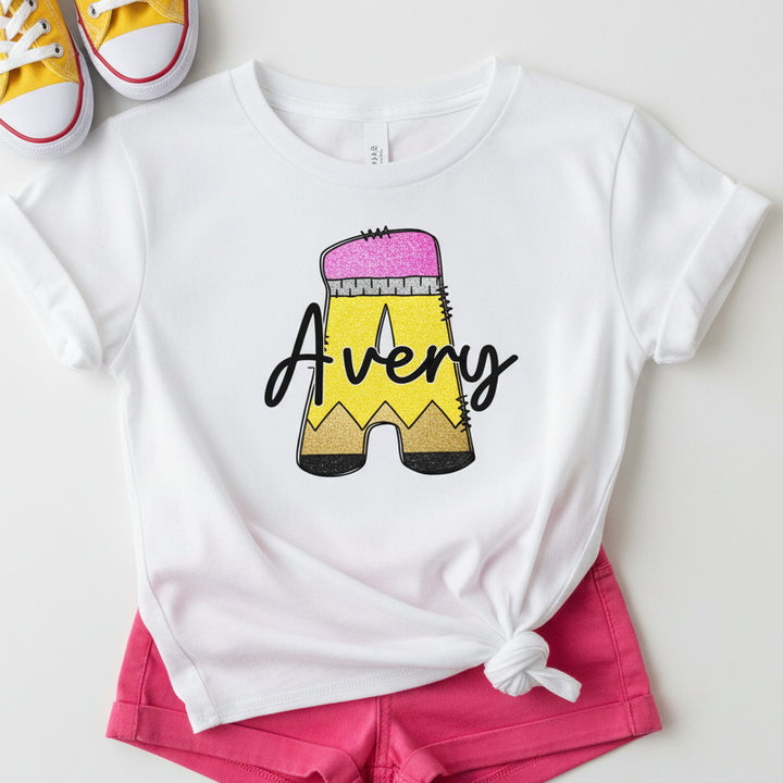 A white short-sleeve T-shirt laid flat with a tied hem, featuring a large pencil graphic and the name 'Avery' in black script. The shirt is styled with bright pink shorts and yellow sneakers on a light neutral background.