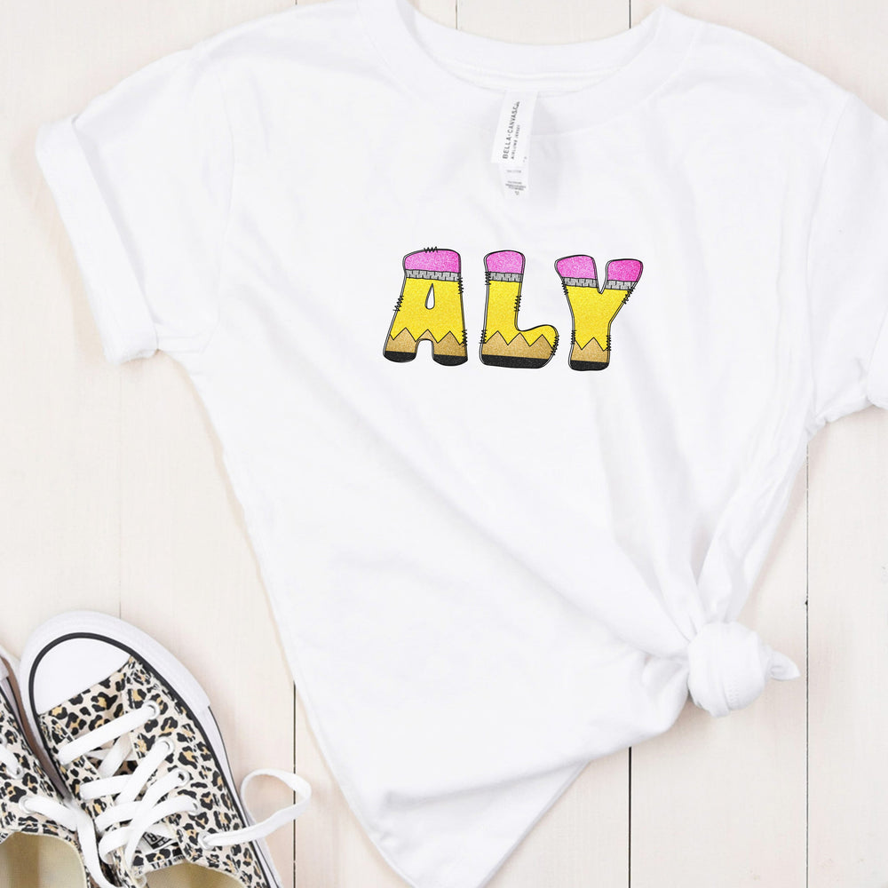 A white short-sleeve t-shirt with pencil letters graphic spelling 'ALY', styled in a flat lay on a light wood background next to leopard print sneakers.