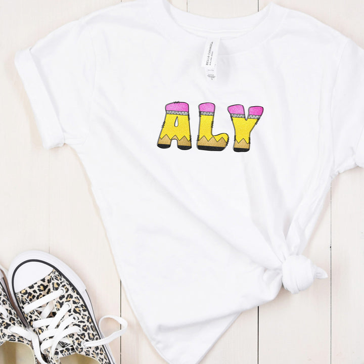 A white short-sleeve t-shirt with pencil letters graphic spelling 'ALY', styled in a flat lay on a light wood background next to leopard print sneakers.