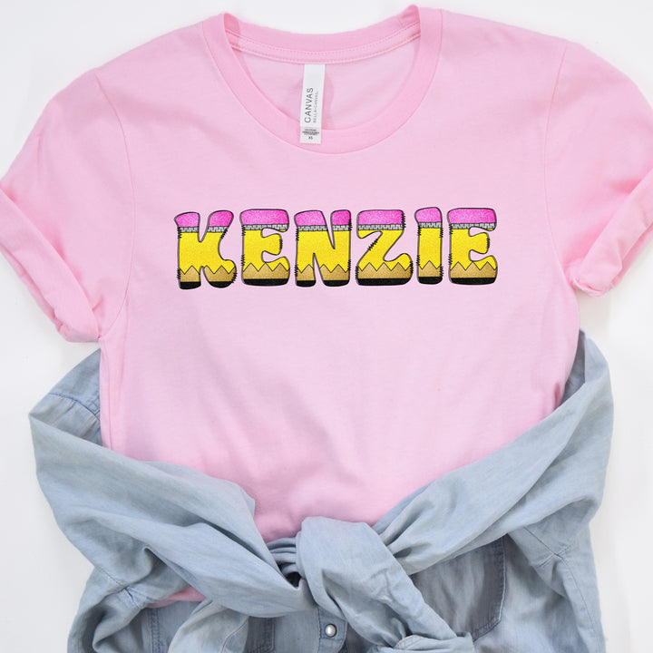 A pink short-sleeve T-shirt with the word 'KENZIE' printed across the chest in a yellow and pink bubbly pencil font, styled flat-lay with a light blue denim shirt tied underneath on a white background.