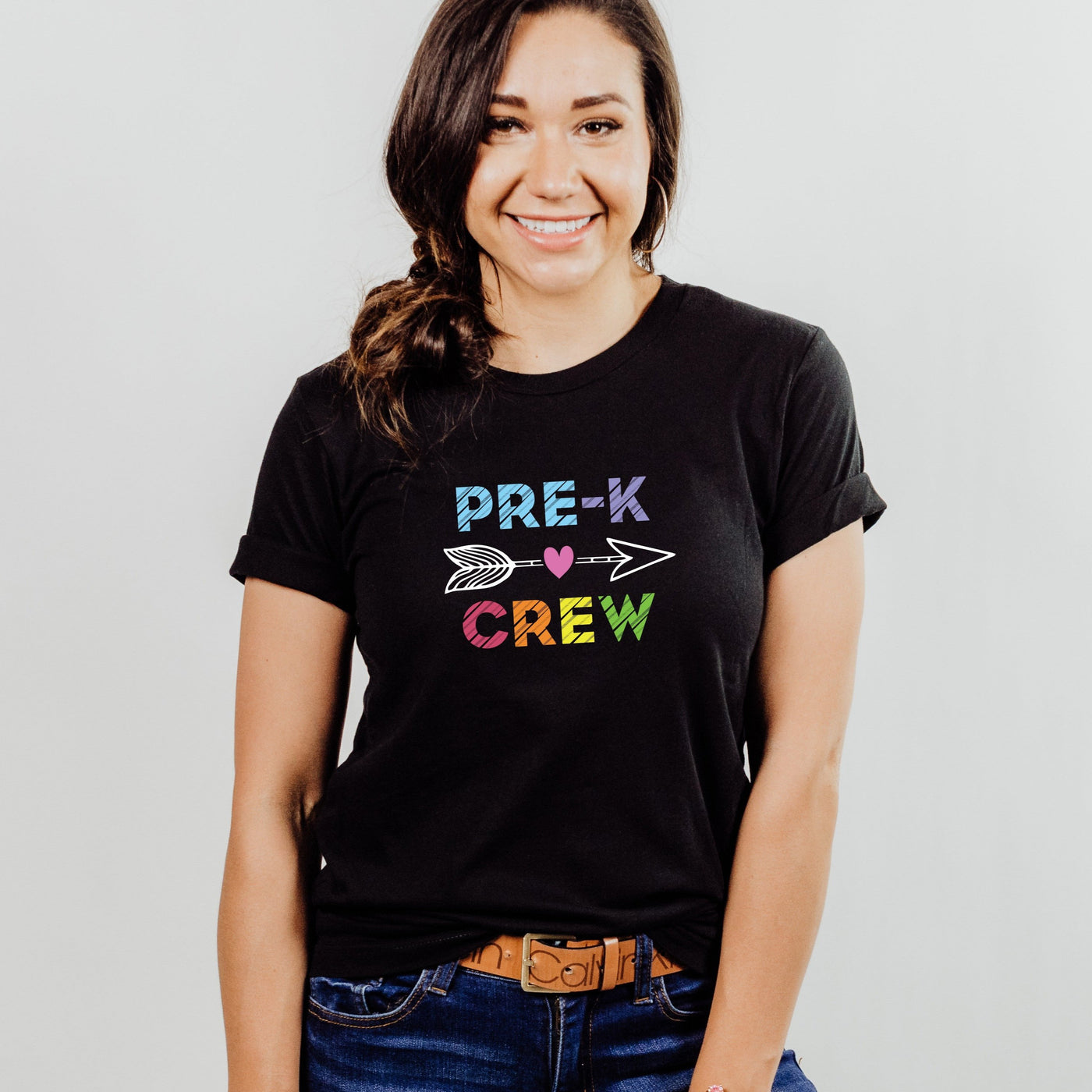 Woman wearing black personalized pre k crew shirt for preschool teachers featuring vibrant, rainbow colored letters spelling Pre-K Crew that pop against the dark fabric, creating a fun and playful look. An eye-catching arrow between the words adds an extra touch of whimsy, making this shirt a standout choice for any preschool teacher.  