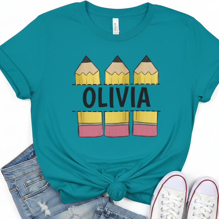 A teal short-sleeve graphic T-shirt laid flat with a knot at the hem, styled with denim shorts and white sneakers. The shirt features a pencil-themed graphic with the name 'OLIVIA' in black across the center.