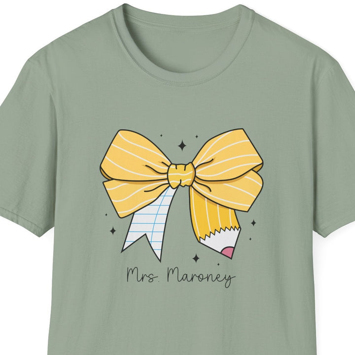 A sage green short-sleeve t-shirt with a graphic of a yellow striped bow, a pencil, and the text 'Mrs. Maroney' printed on the front, shown against a plain white background.