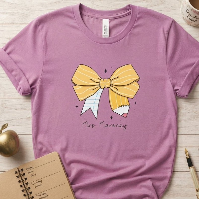 A lavender-purple short-sleeve t-shirt with a yellow and white pencil-themed bow graphic and the text 'Mrs. Maroney' printed on the front.
