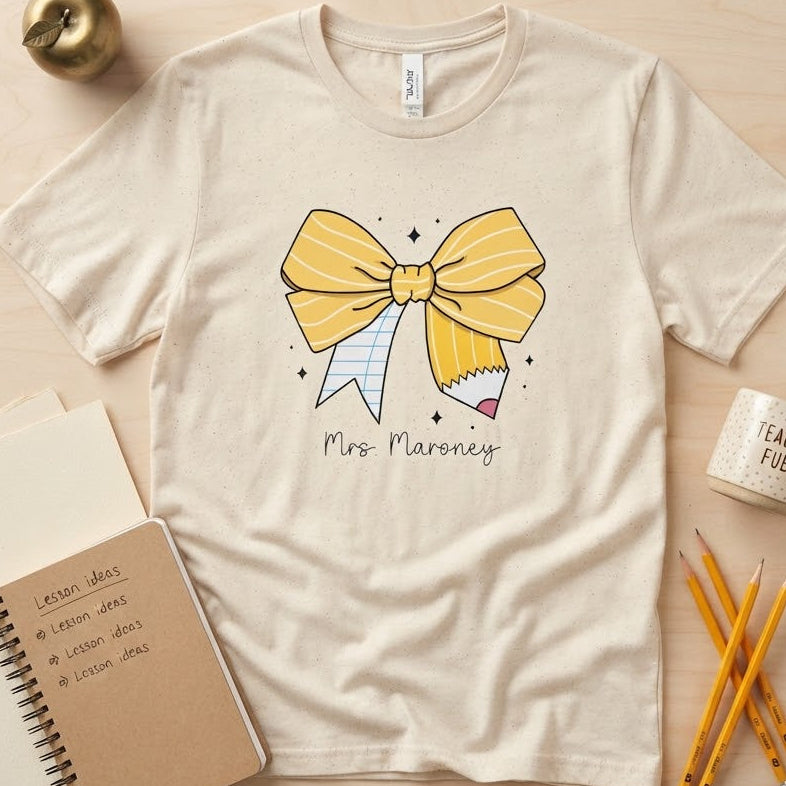 A cream-colored short-sleeve t-shirt with a graphic of a yellow bow holding a pencil, with the text 'Mrs. Maroney' printed below.