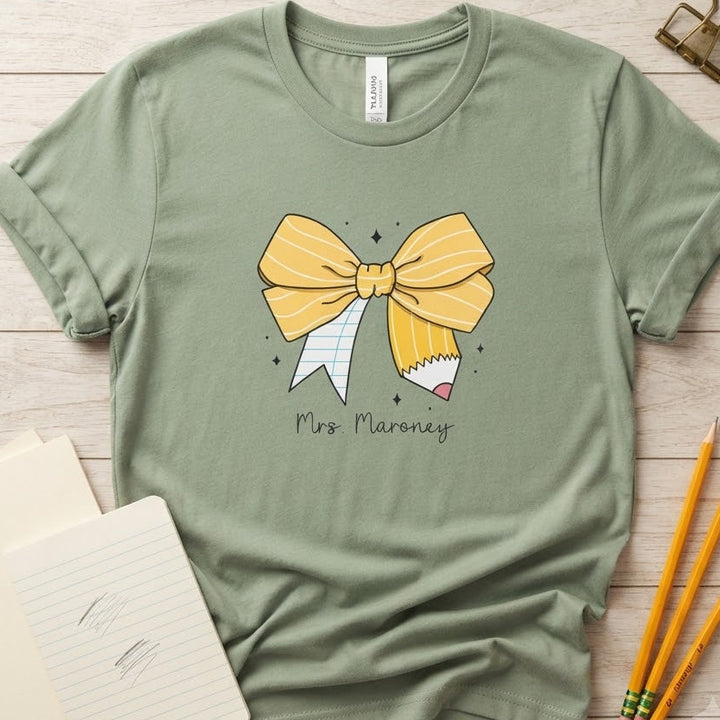 A sage green short-sleeve t-shirt with a graphic of a yellow bow made from a pencil and ribbon, with the text 'Mrs. Maroney' printed below.