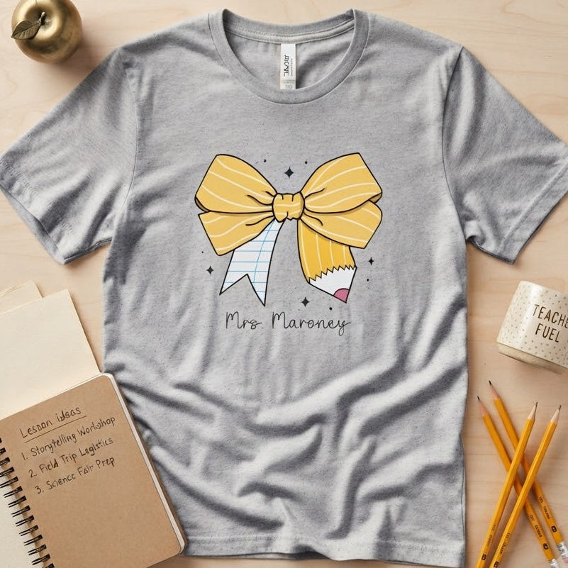 A light gray short-sleeve t-shirt with a yellow and white bow graphic and the text 'Mrs. Maroney' printed on the front, styled in a flat lay on a light wood background with a notebook, pencil, mug, and small decorative object.