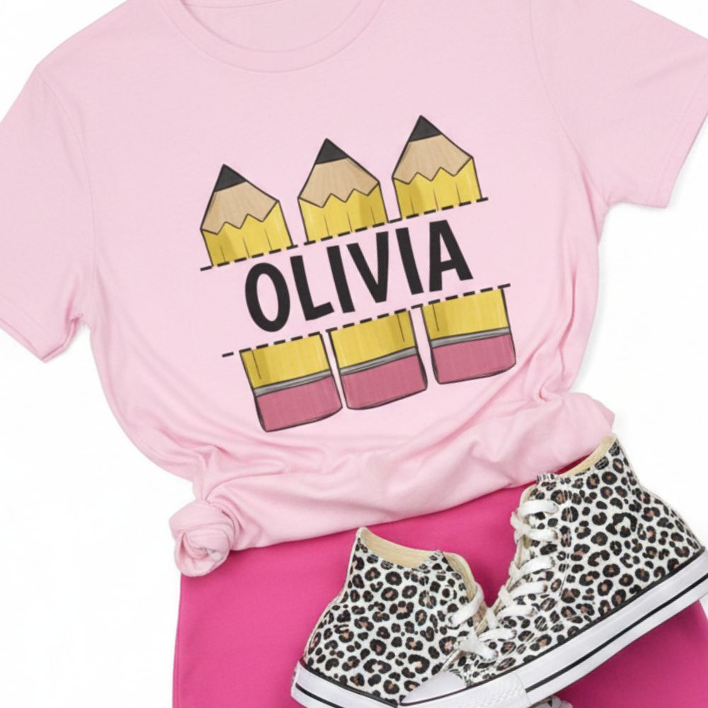 A pink short-sleeve T-shirt with a graphic of three yellow-and-pink pencils and the name 'OLIVIA' in black across the chest, styled in a flat lay with hot pink bottoms and leopard-print high-top sneakers on a white background.