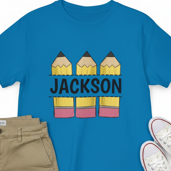 A bright blue short-sleeve T-shirt laid flat with a graphic of three yellow pencils above and below the name 'JACKSON' in black. Folded khaki pants and white sneakers are partially visible around the shirt as styling props on a white background.
