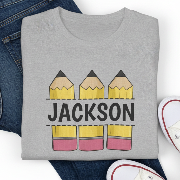 A heather gray short-sleeve T-shirt laid flat with a graphic of three yellow pencils above and below the name 'JACKSON' in black. Folded jeans and white sneakers are partially visible around the shirt as styling props on a white background.