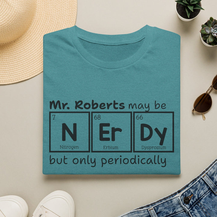 Folded teal blue t-shirt with black text graphic that reads 'Mr. Roberts may be nerdy but only periodically' with nerdy spelled out in periodic elements. surrounded by a hat, sunglasses, shoes, and plants on a light surface.