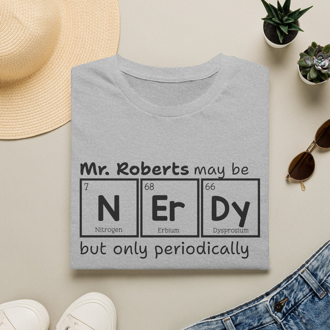 A folded light gray T-shirt with a black printed graphic reading 'Mr. Roberts may be NEr Dy but only periodically' styled in a flat lay with a straw hat, small potted succulents, sunglasses, white sneakers, and blue jeans on a light beige background.