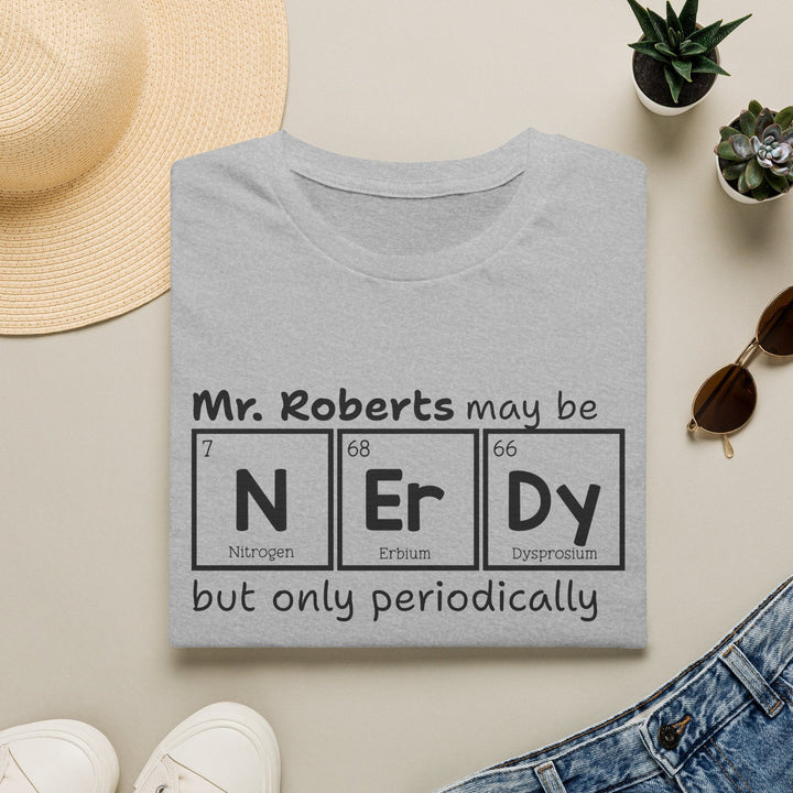 A folded light gray T-shirt with a black printed graphic reading 'Mr. Roberts may be NEr Dy but only periodically' styled in a flat lay with a straw hat, small potted succulents, sunglasses, white sneakers, and blue jeans on a light beige background.