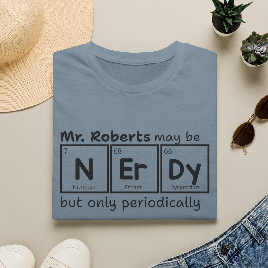 Folded blue t-shirt with black text graphic that reads 'Mr. Roberts may be nerdy but only periodically' with nerdy spelled out in periodic elements. surrounded by a hat, sunglasses, shoes, and plants on a light surface.