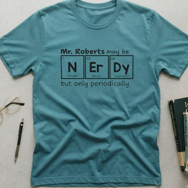 A teal blue graphic t-shirt laid flat on a light gray textured surface, surrounded by props including eyeglasses, a pen, and books. The shirt features a black printed graphic that reads 'Mr. Roberts may be nerdy but only periodically' with nerdy spelled out in periodic table elements.