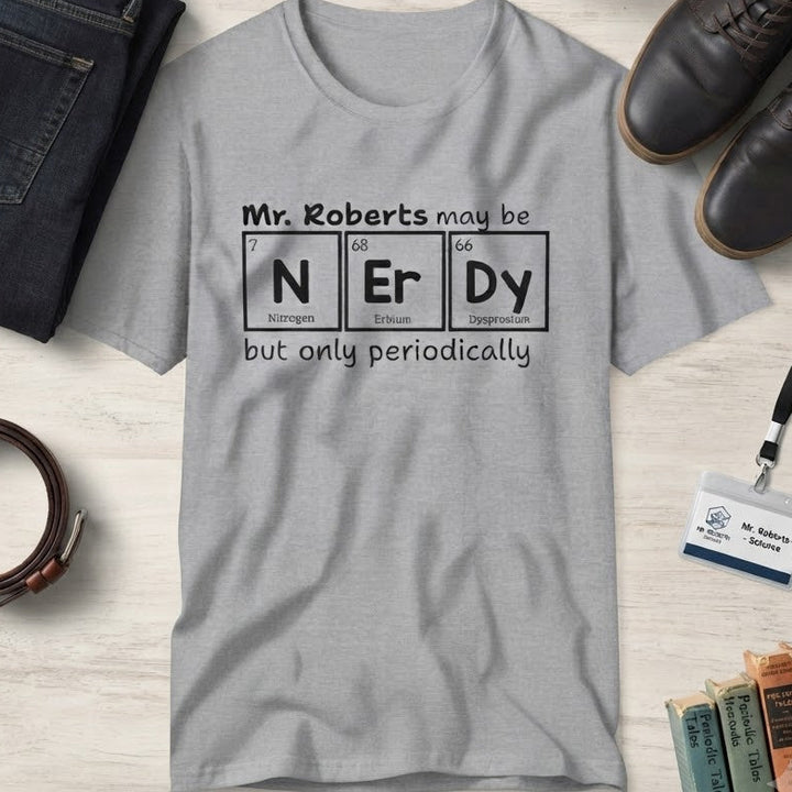 A light gray graphic t-shirt laid flat on a light wood surface, surrounded by jeans, shoes, a belt, pencils, books, and a small plant. The shirt has a black text graphic that reads 'Mr. Roberts may be nerdy but only periodically' with nerdy spelled out in periodic elements.