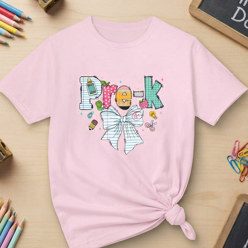 A light pink short-sleeve t-shirt with a graphic design spelling 'Pre-K' using school-themed items and a preppy bow styled in a flat-lay on a light wood desk with pencils, sharpeners, paper clips, and chalkboard signs.