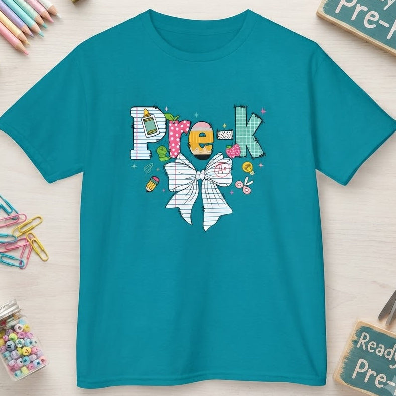 A teal short-sleeve t-shirt with a colorful 'Pre-k' graphic and white bow design, presented in a flat-lay arrangement with school-themed props on a light wood background.