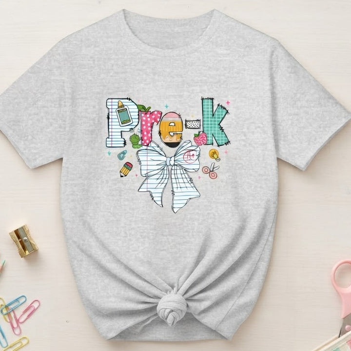 A light ash gray t-shirt with a knotted hem, featuring a colorful graphic on the front that spells 'Pre-K' with a bow tie and cartoon details. The t-shirt is styled in a flat-lay arrangement on a light wood background with school supplies including colored pencils, paper clips, a pencil case, and scissors.