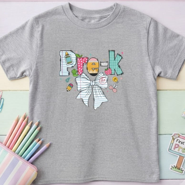 A heather gray short-sleeve t-shirt with a colorful graphic spelling 'Pre-K' using candy and a bow tie, styled in a pastel flat-lay with stationery props.