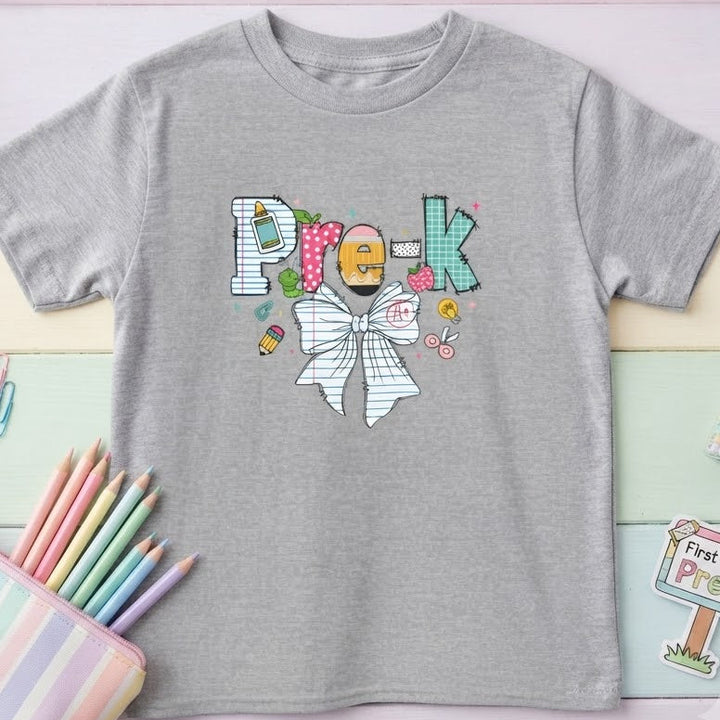 A heather gray short-sleeve t-shirt with a colorful graphic spelling 'Pre-K' using candy and a bow tie, styled in a pastel flat-lay with stationery props.