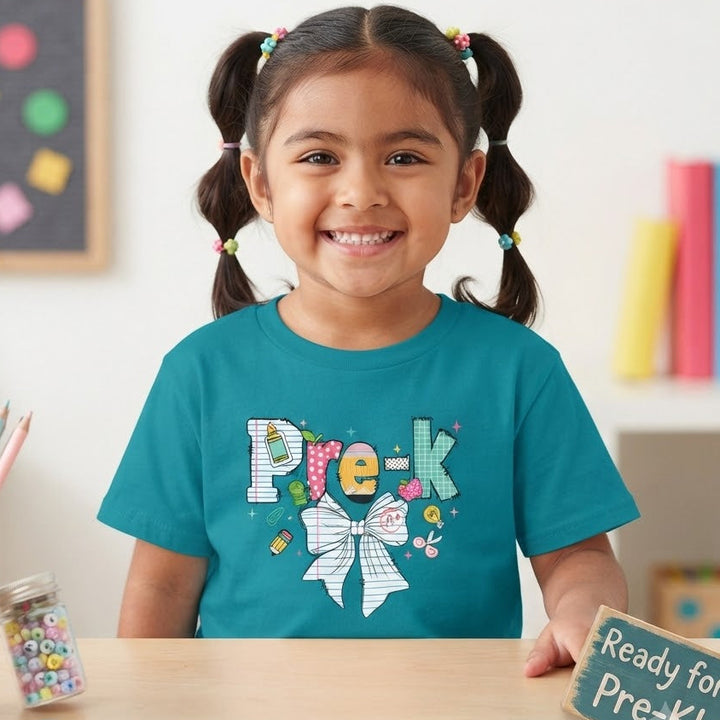 A young girl with dark hair in pigtails smiles while wearing a teal graphic t-shirt that says 'Pre-K' with a bow design. She is sitting at a wooden desk with school supplies, including a jar of colorful candies, a cup of colored pencils, and pink scissors, with a small chalkboard sign reading 'Ready for Pre-K!' in a bright classroom setting.
