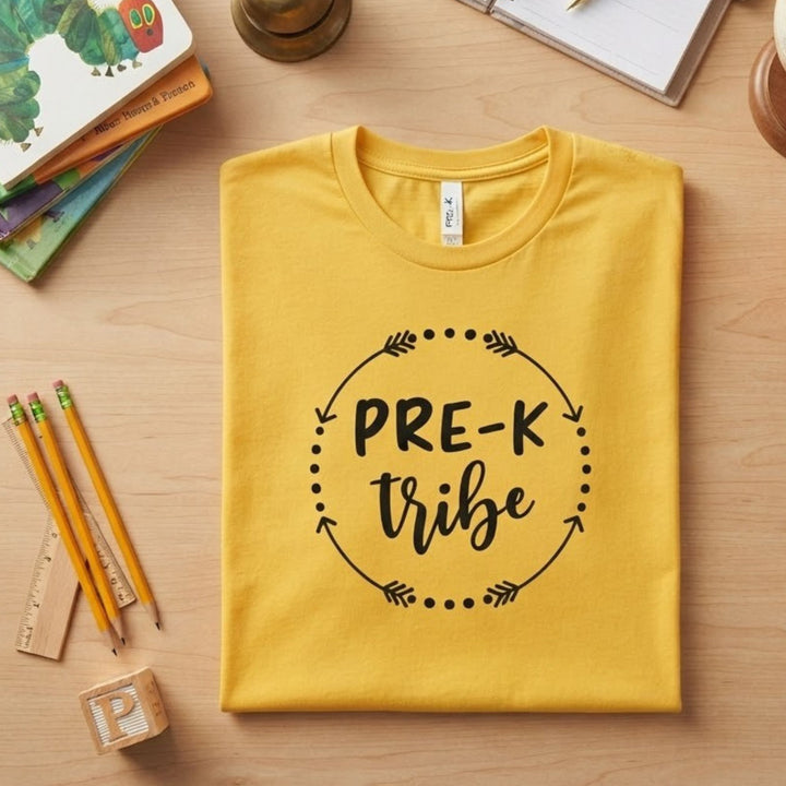A folded yellow t-shirt with a black graphic reading 'PRE-K tribe' inside a dotted circle with arrows, displayed on a light wood desk with school supplies.