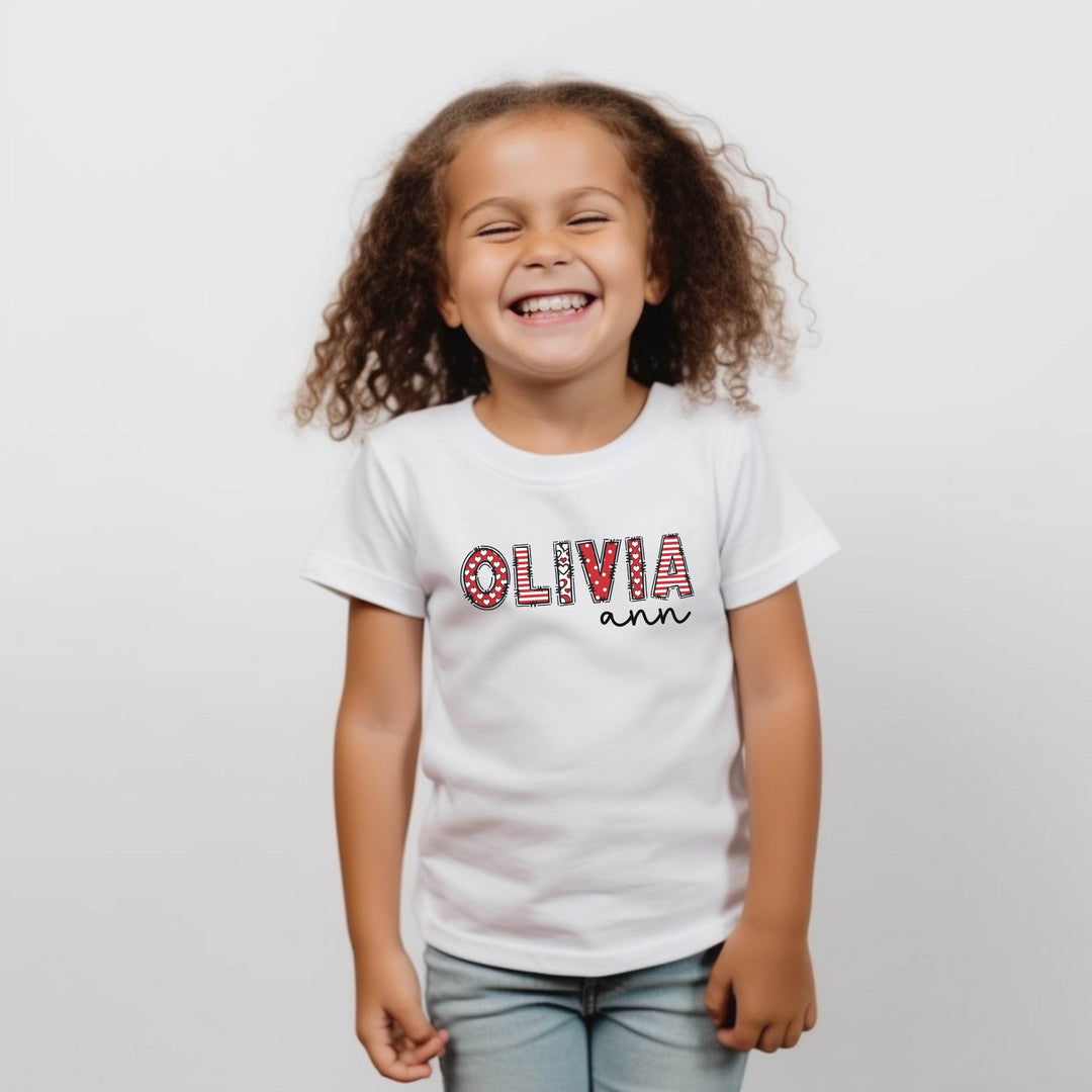 Child wearing a white t-shirt with 'OLIVIA ann' printed on it against a plain background. First name letters are combination of red and white stripes and polka dots. Other name is in black modern script.