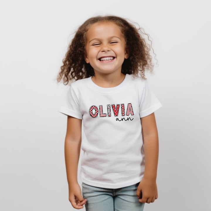 Child wearing a white t-shirt with 'OLIVIA ann' printed on it against a plain background. First name letters are combination of red and white stripes and polka dots. Other name is in black modern script.