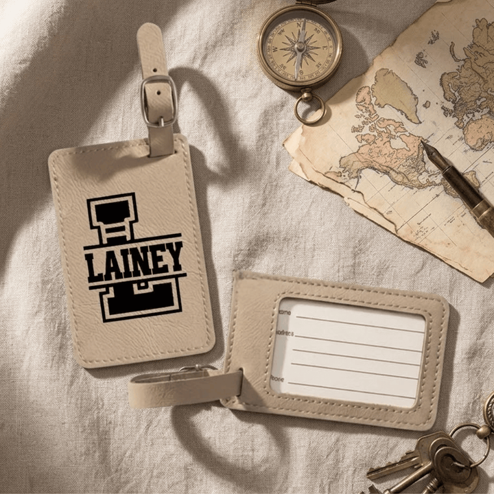 Two beige luggage tags on a textured surface with a world map and compass in the background. Front has a varsity style split monogram engraved in black. The back shows a clear plastic opening with a removable card for personal information.