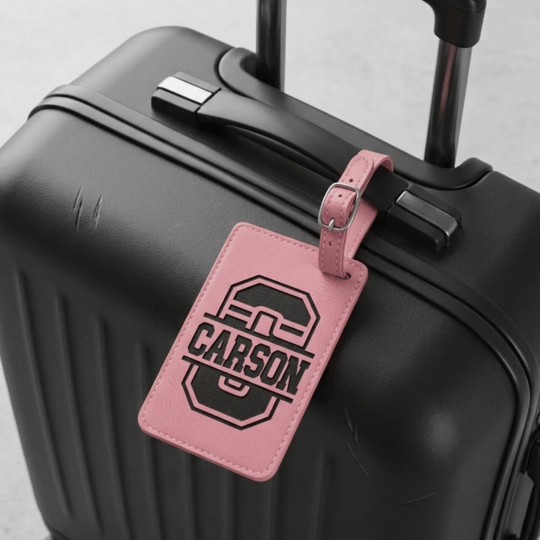 Black suitcase with a pink luggage tag featuring 'Carson' engraved in black in a varsity font.