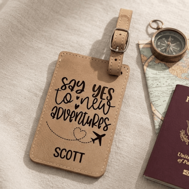 A personalized tan leather luggage tag with black script text engraved reading 'say yes to new adventures' and the name 'SCOTT', styled on a light fabric surface with a compass, map, and passport nearby.