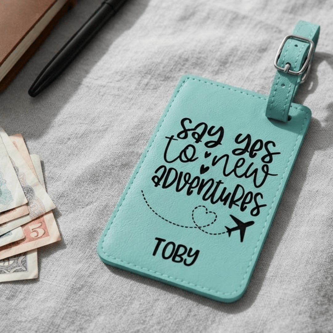 A personalized teal faux-leather luggage tag with a silver buckle, featuring the engraved black text 'say yes to new ADVENTURES' and the name 'TOBY'. The tag is displayed on a textured gray fabric surface alongside a pen, a notebook, and some banknotes.