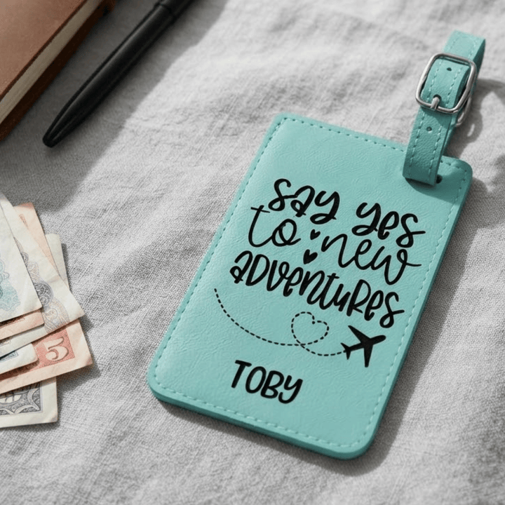 A personalized teal faux-leather luggage tag with a silver buckle, featuring the engraved black text 'say yes to new ADVENTURES' and the name 'TOBY'. The tag is displayed on a textured gray fabric surface alongside a pen, a notebook, and some banknotes.