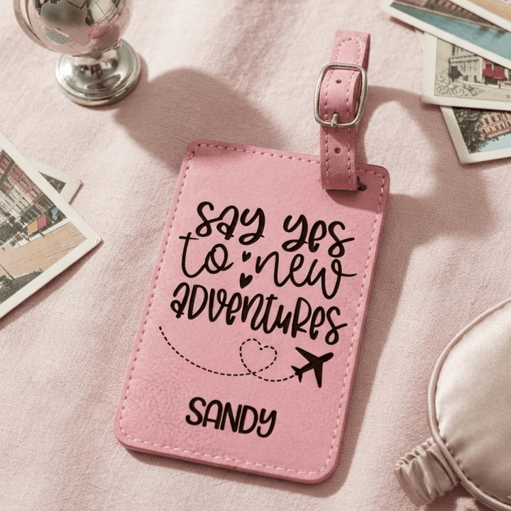 A pink faux-leather luggage tag with a silver buckle, featuring the black engraved text 'say yes to new adventures' and the name 'SANDY' with a small heart and airplane graphic. The tag is styled on a pink fabric surface with travel-themed props.