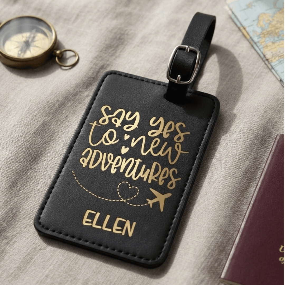 A black faux-leather luggage tag with gold metallic engraved lettering that reads 'say yes to new adventures' and the name 'ELLEN', shown on a textured beige fabric surface with a compass, map, and passport nearby.