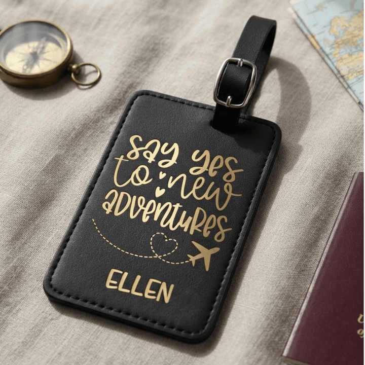A black faux-leather luggage tag with gold metallic engraved lettering that reads 'say yes to new adventures' and the name 'ELLEN', shown on a textured beige fabric surface with a compass, map, and passport nearby.