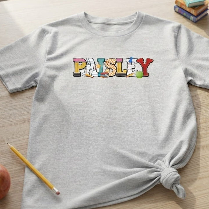A light gray heather t-shirt with the name 'PAISLEY' printed across the chest in colorful, playful school letters. The t-shirt is styled in a flat-lay arrangement on a light wood desk, surrounded by school-themed props including a pencil, apple, scissors, books, and wooden blocks.