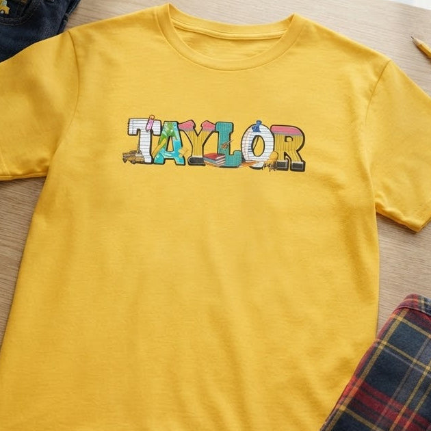 A yellow short-sleeve T-shirt laid flat on a wooden table, with the word "TAYLOR" printed across the chest in multicolored school letters. Surrounding props include folded dark jeans, a plaid shirt, striped socks, a pencil, and a small notebook near a window.