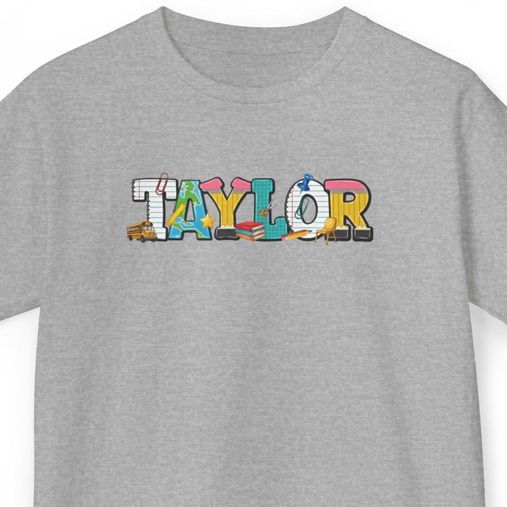 A heather gray short-sleeve t-shirt with the word 'TAYLOR' printed across the chest in a colorful, patterned design featuring small school supply illustrations.